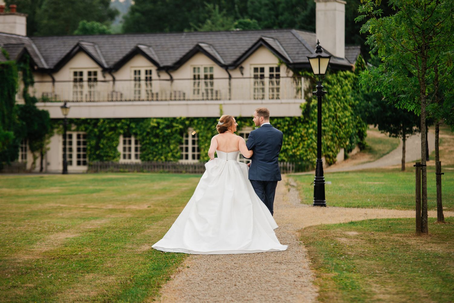 A couple in wedding attire walking down a gravel path toward a stately manor house with dormer windows and lush greenery.
