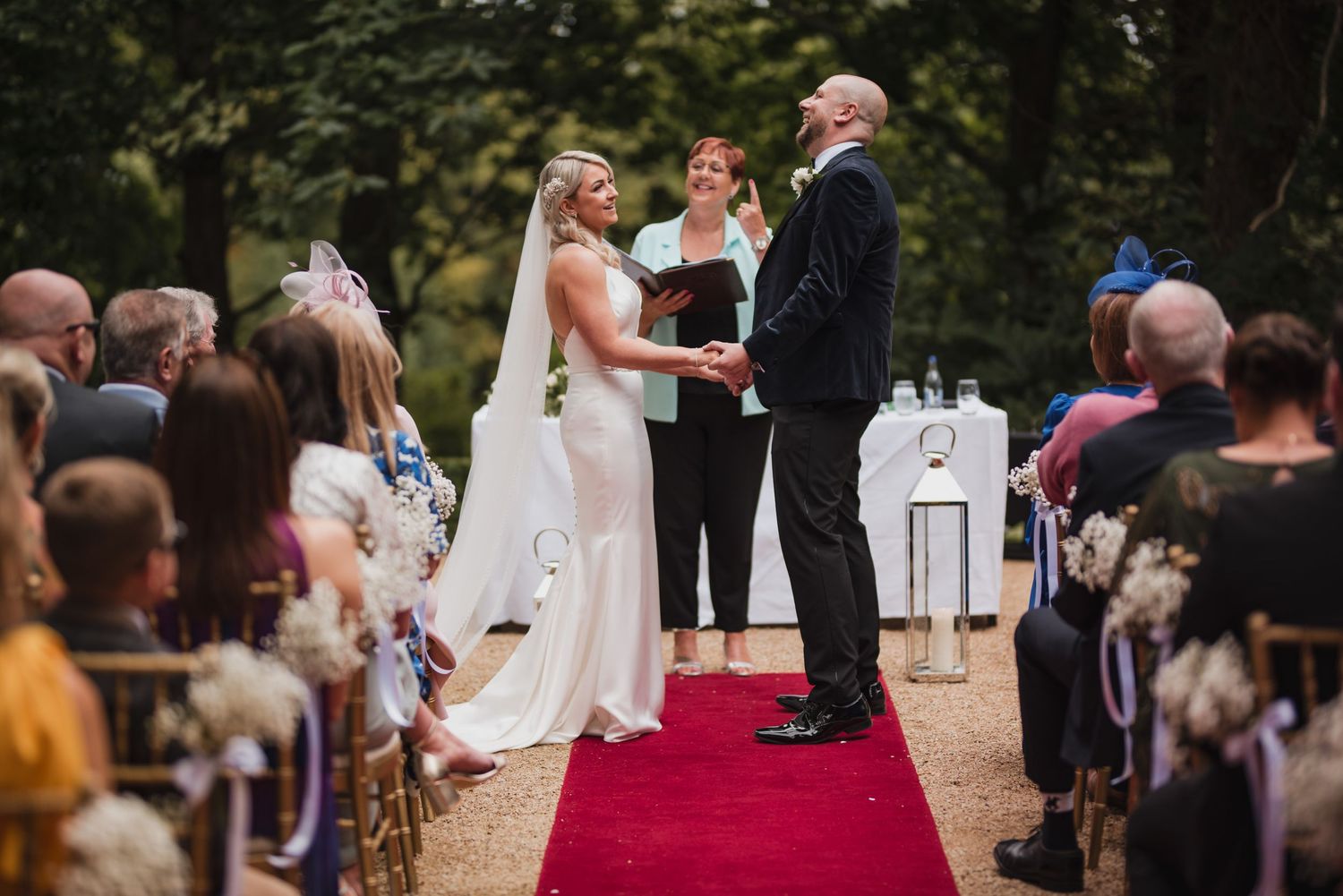 A couple exchanges vows during an outdoor forest wedding ceremony on a red carpet with guests seated on both sides.