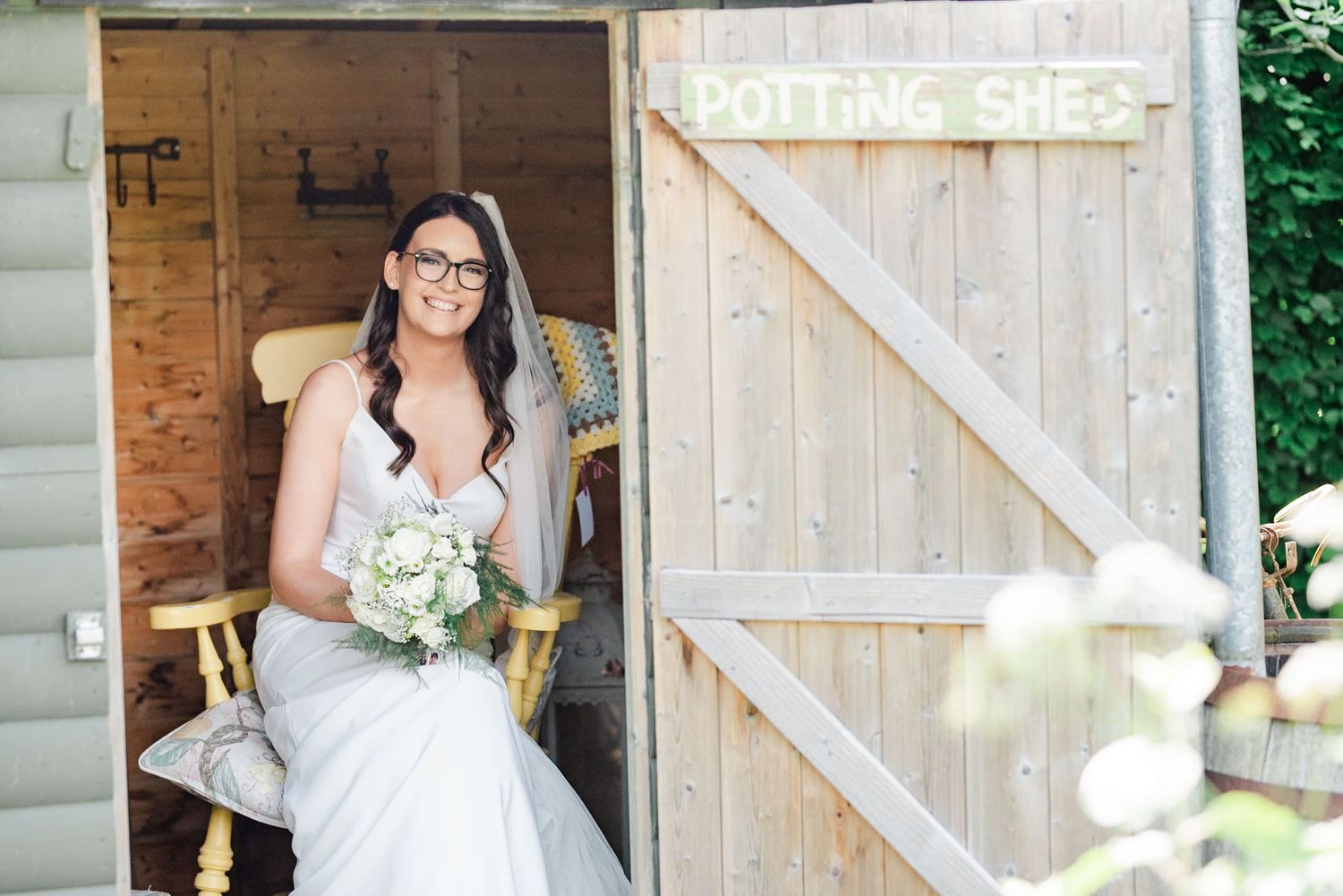 Bride in wedding dress sits in rustic wooden barn doorway holding white bouquet.