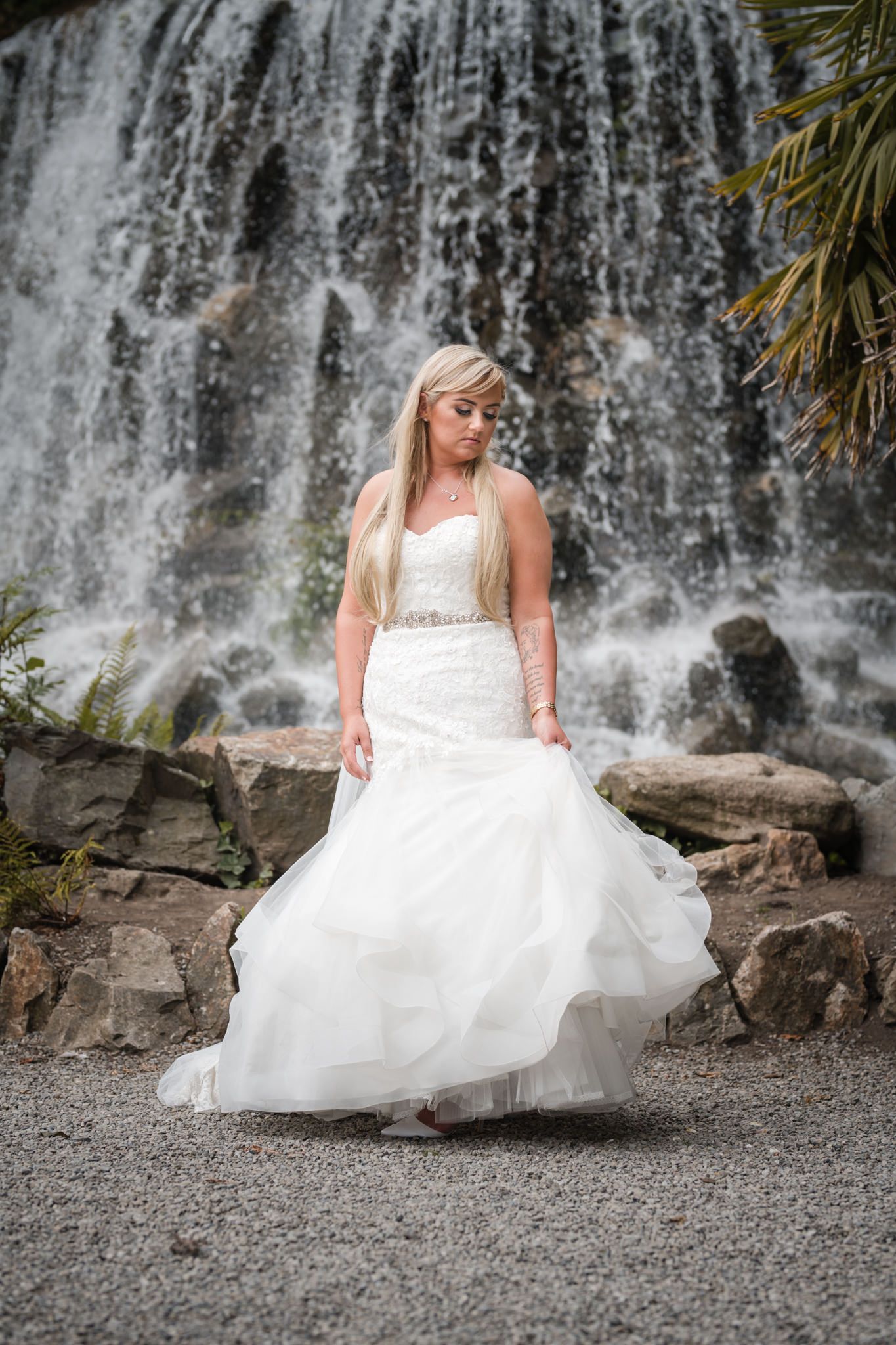A bride in a flowing white wedding dress poses gracefully against a scenic waterfall backdrop.