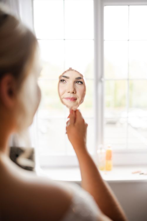A person in a white towel looks into a mirror with a relaxed expression in a bright bathroom setting.