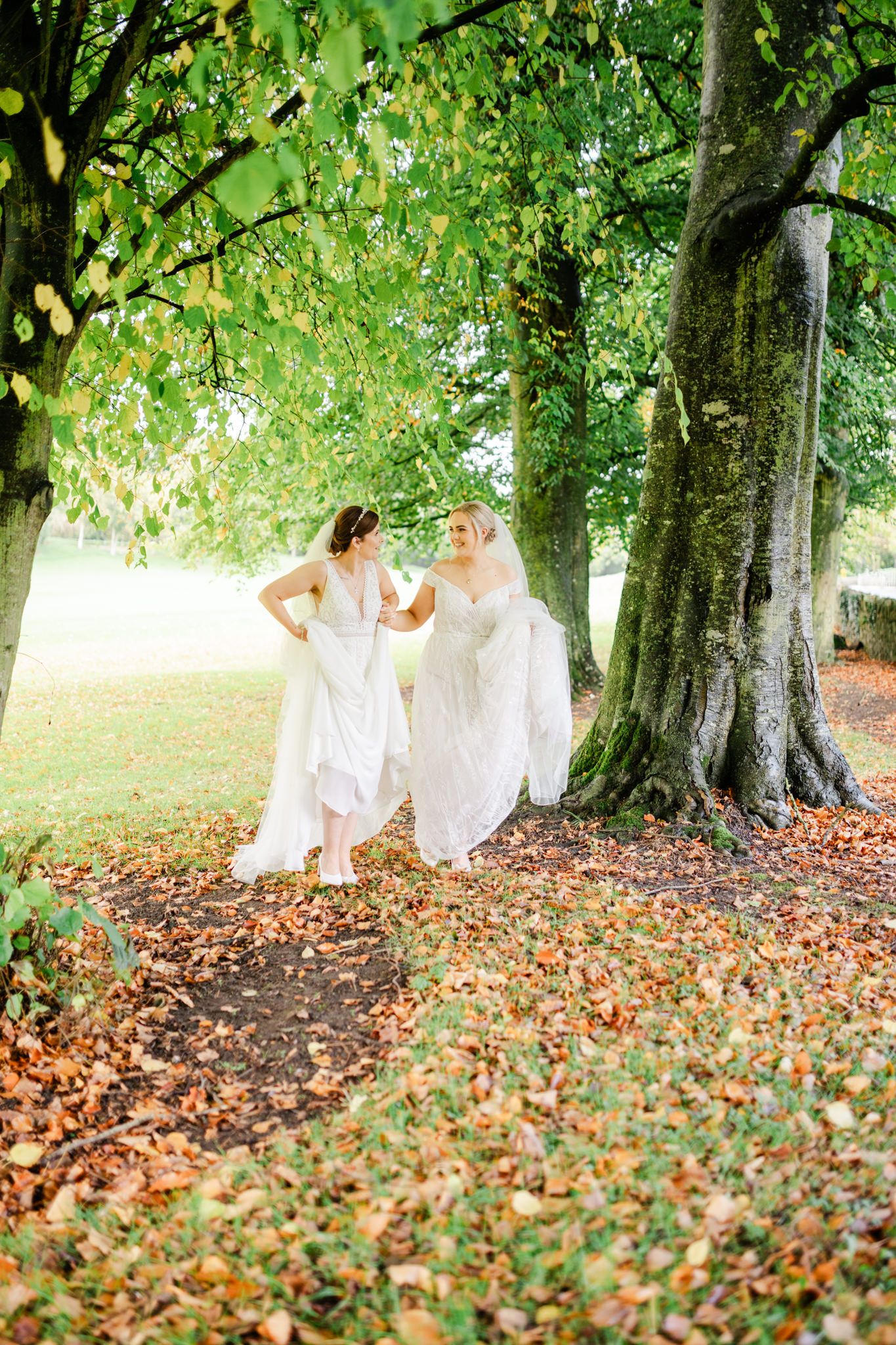 Two brides in white dresses share a joyful moment under green trees with fallen autumn leaves.