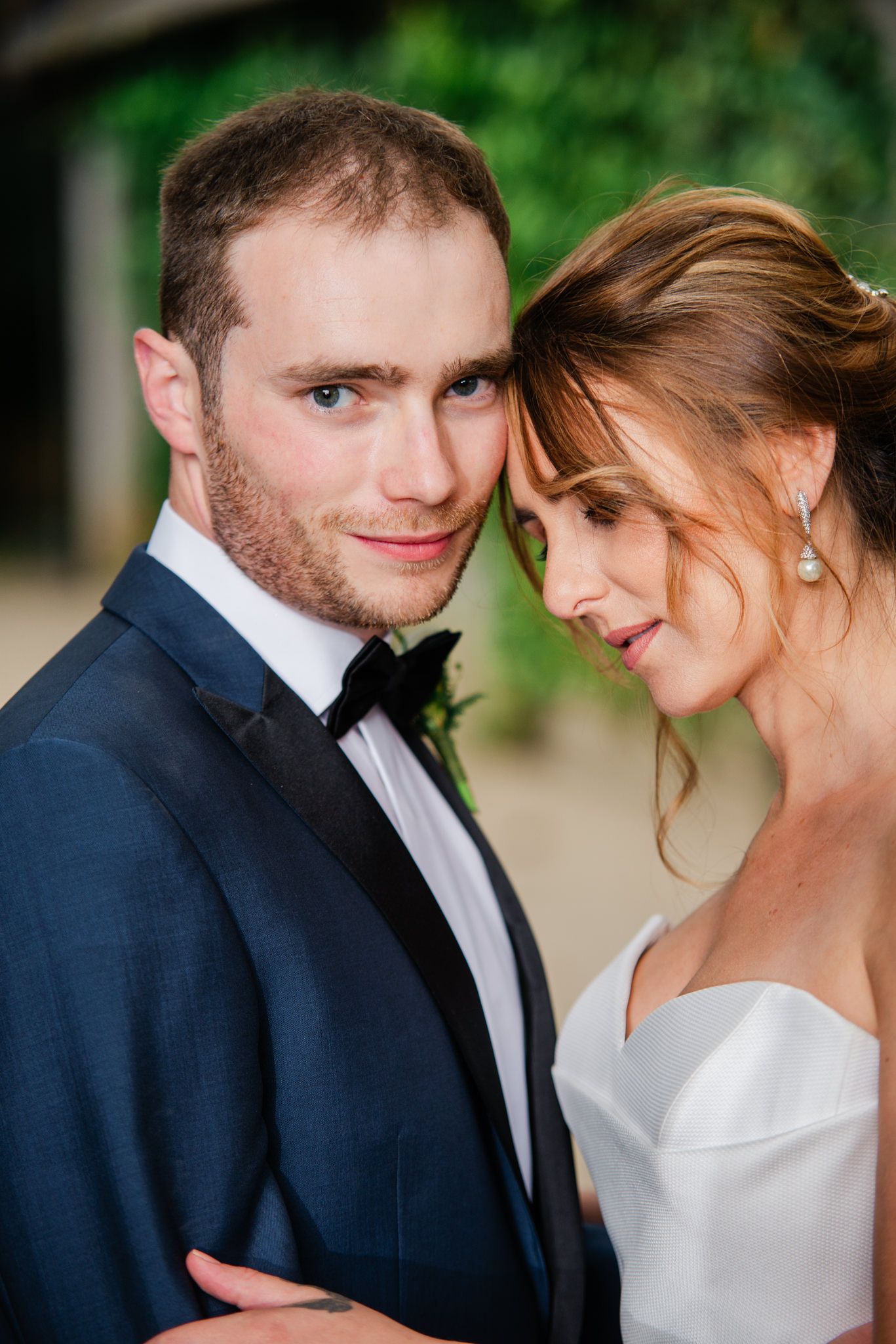 A romantic wedding portrait of a couple in formal attire sharing an intimate moment against a green outdoor background.