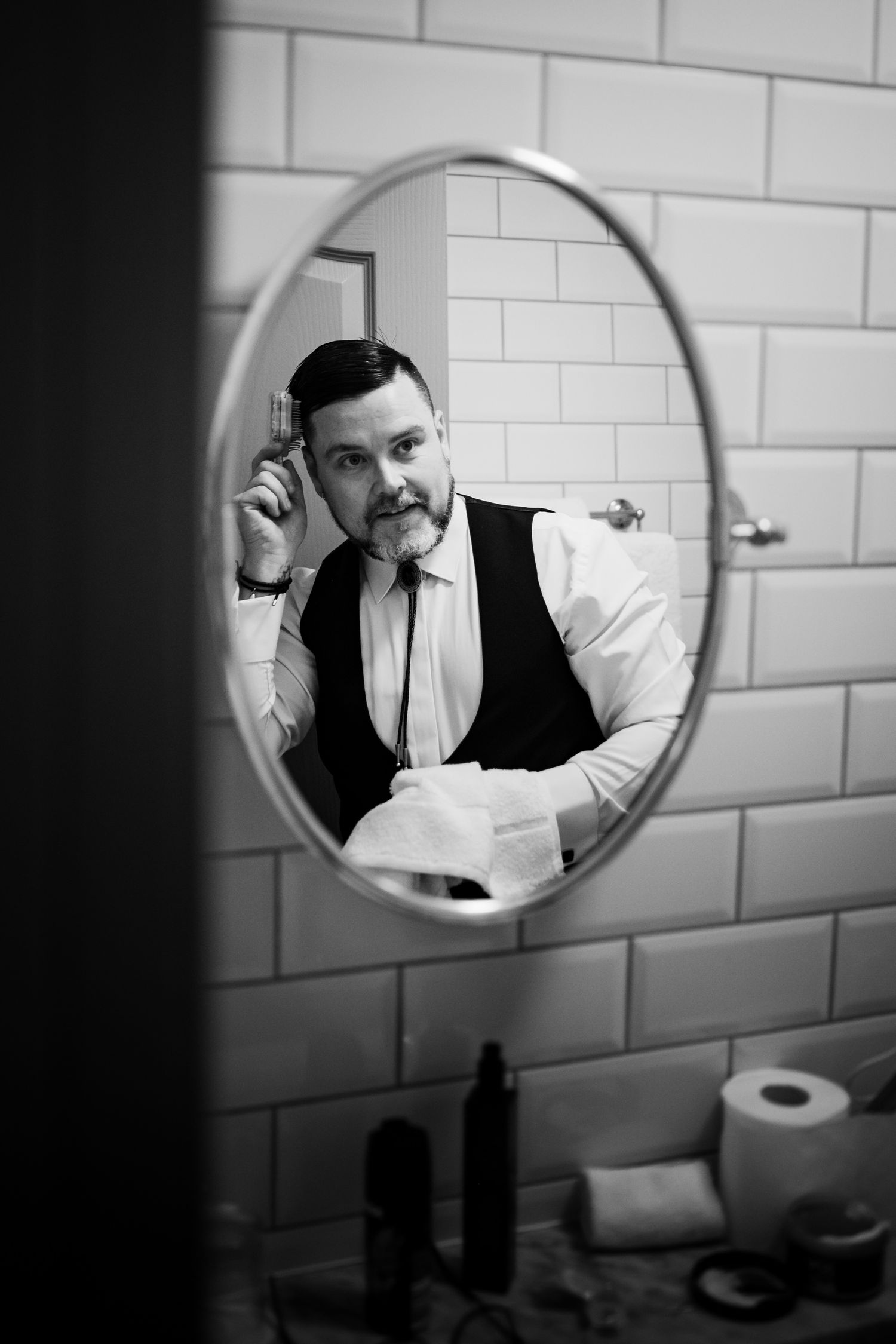 Black and white reflection in circular bathroom mirror shows person adjusting tie while wearing formal vest and shirt.