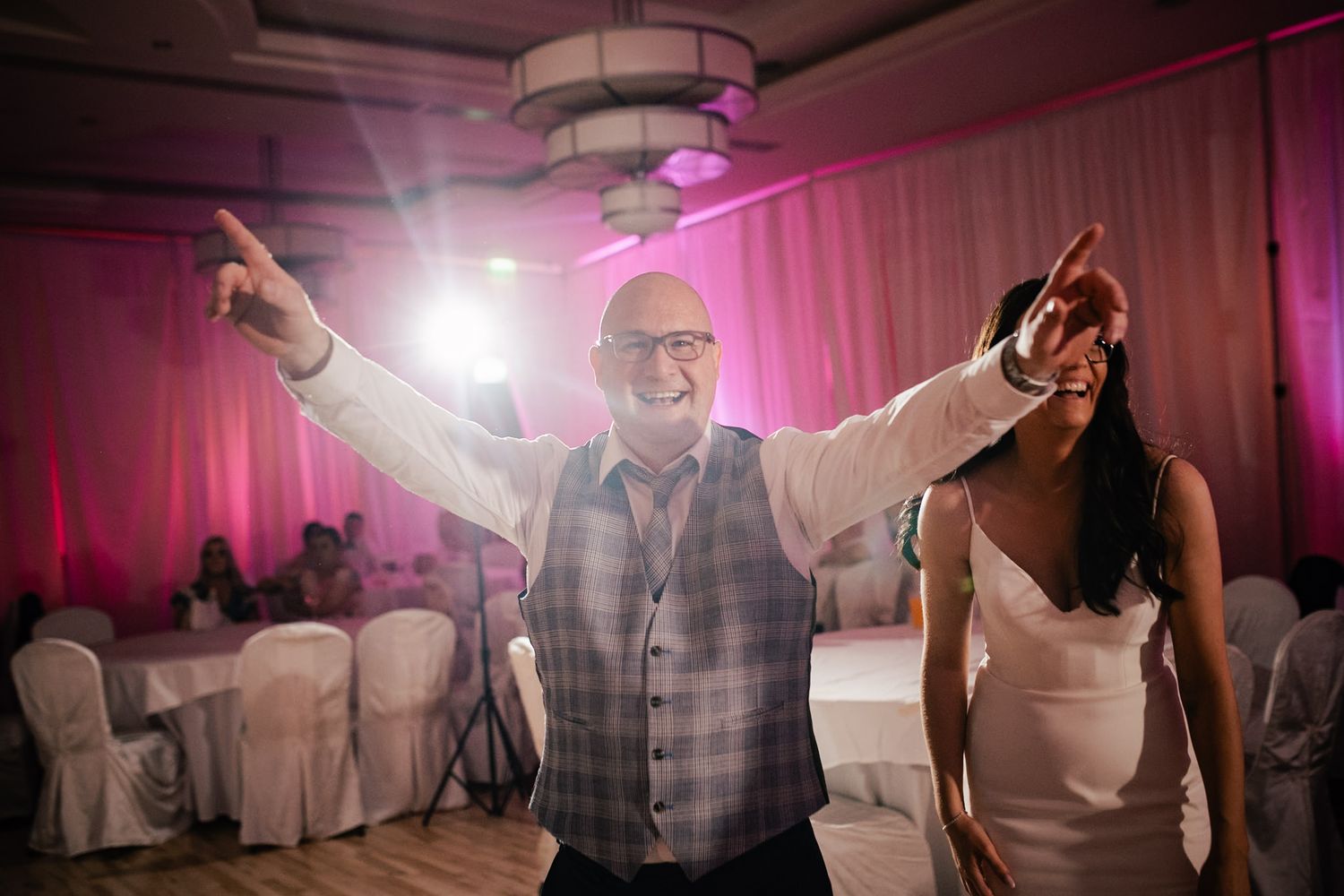 A joyful guest dances with arms raised at a wedding reception with pink uplighting and covered chairs in the background.