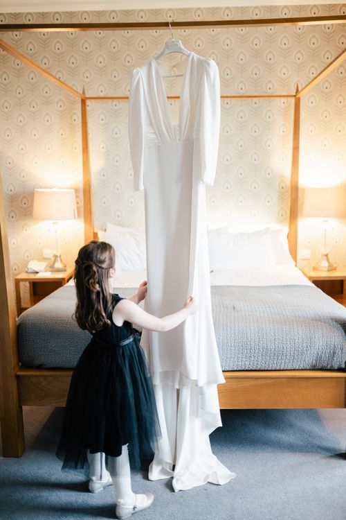 A small figure in a navy tulle dress touches a white wedding gown hanging in front of a canopy bed with illuminated bedside lamps.