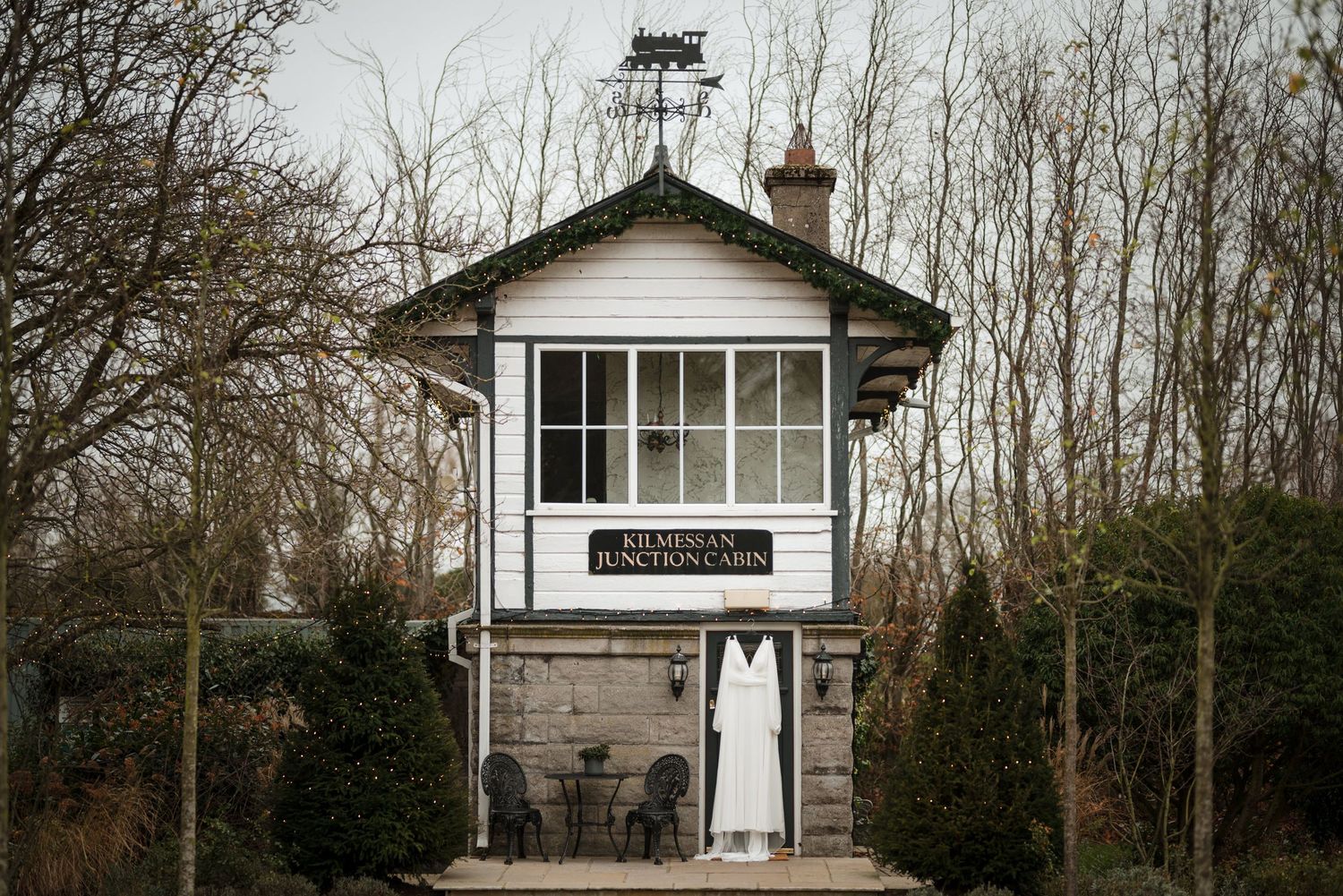 A vintage railway signal box building with white windows and dark trim stands among bare trees on a cloudy day.