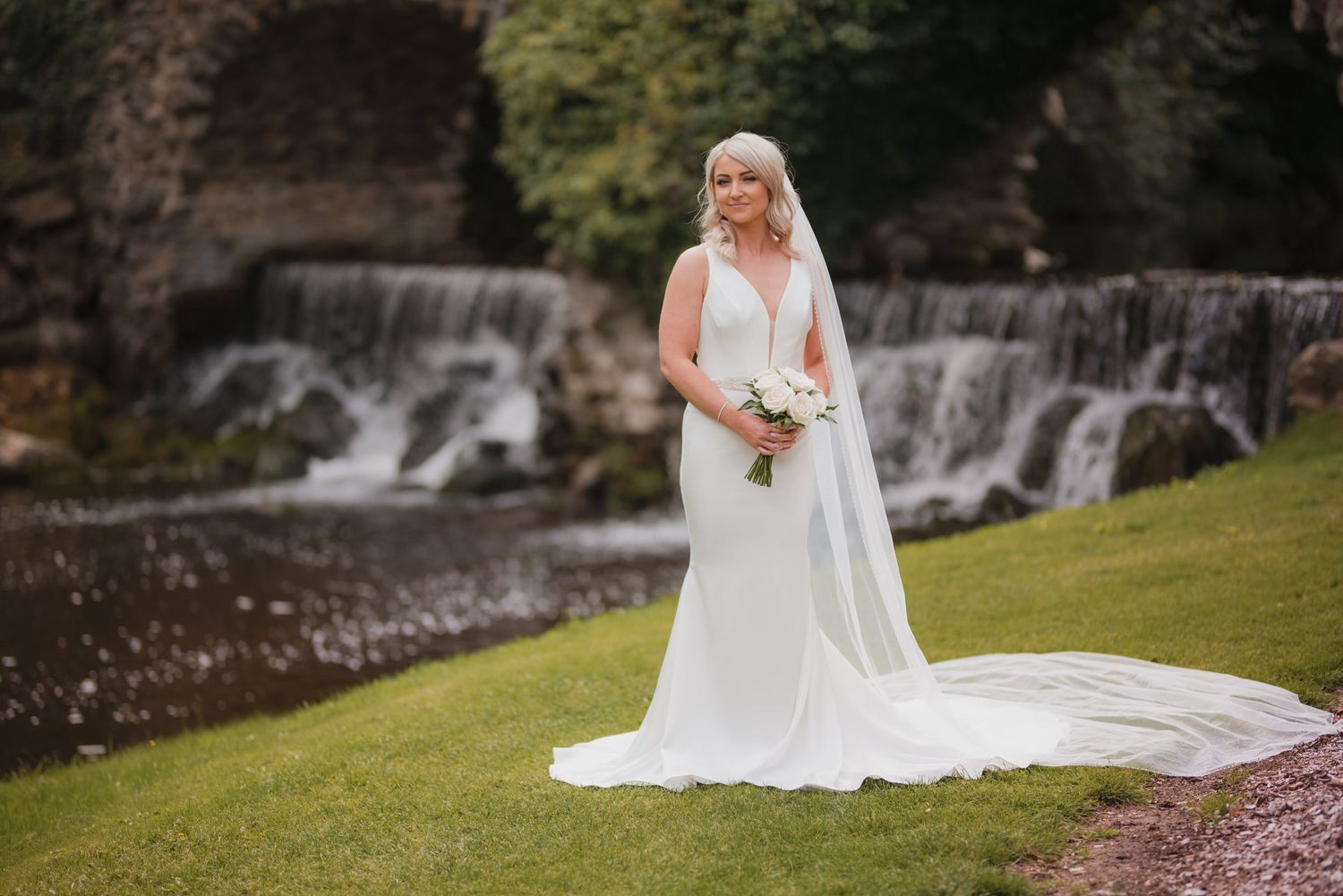 Elegant wedding portrait in white gown against scenic waterfall backdrop in garden setting.