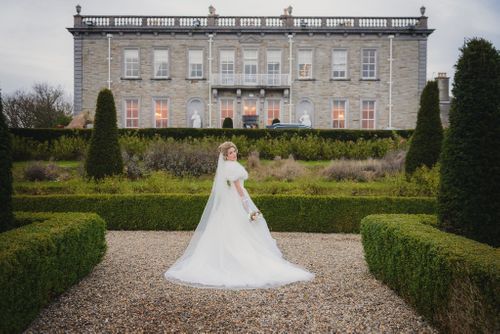Beautiful grand estate with manicured gardens serves as backdrop for elegant white wedding gown in formal garden setting.