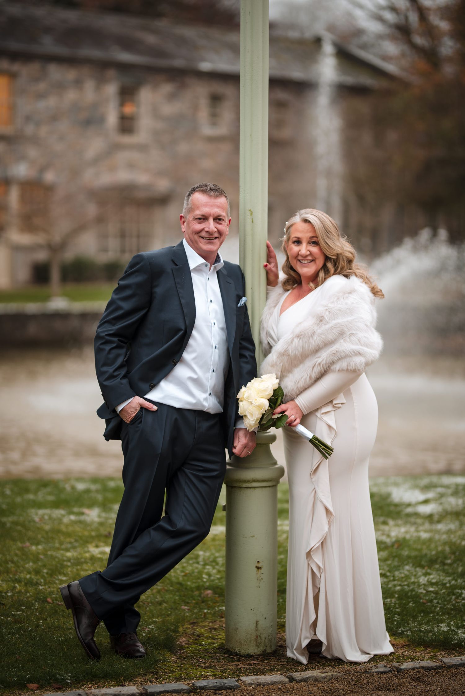 A couple in wedding attire playfully poses with a green pole in a winter garden setting creating fun portrait shots.