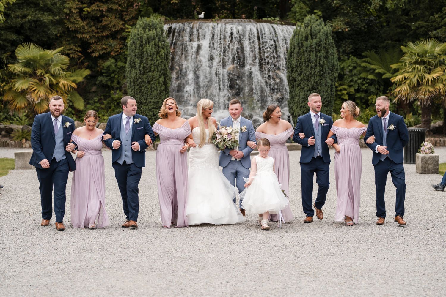 Wedding party in navy suits and blush gowns walks together in a candid shot against a natural outdoor setting.