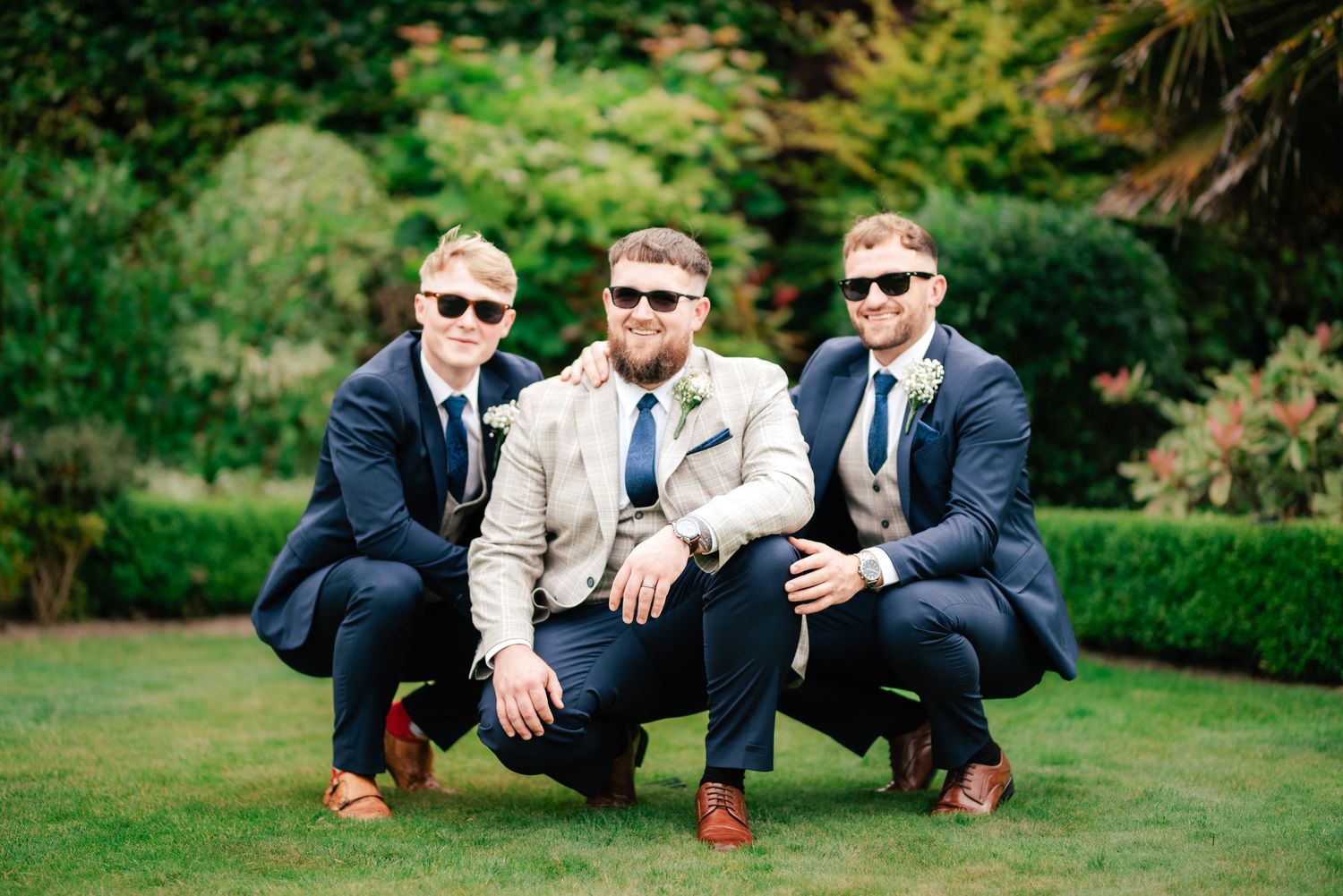 Three groomsmen in sunglasses and suits pose crouching on a manicured garden lawn.
