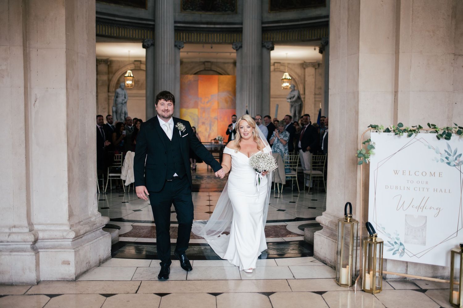 A wedding couple celebrates as they exit a columned venue with lanterns and floral decorations.