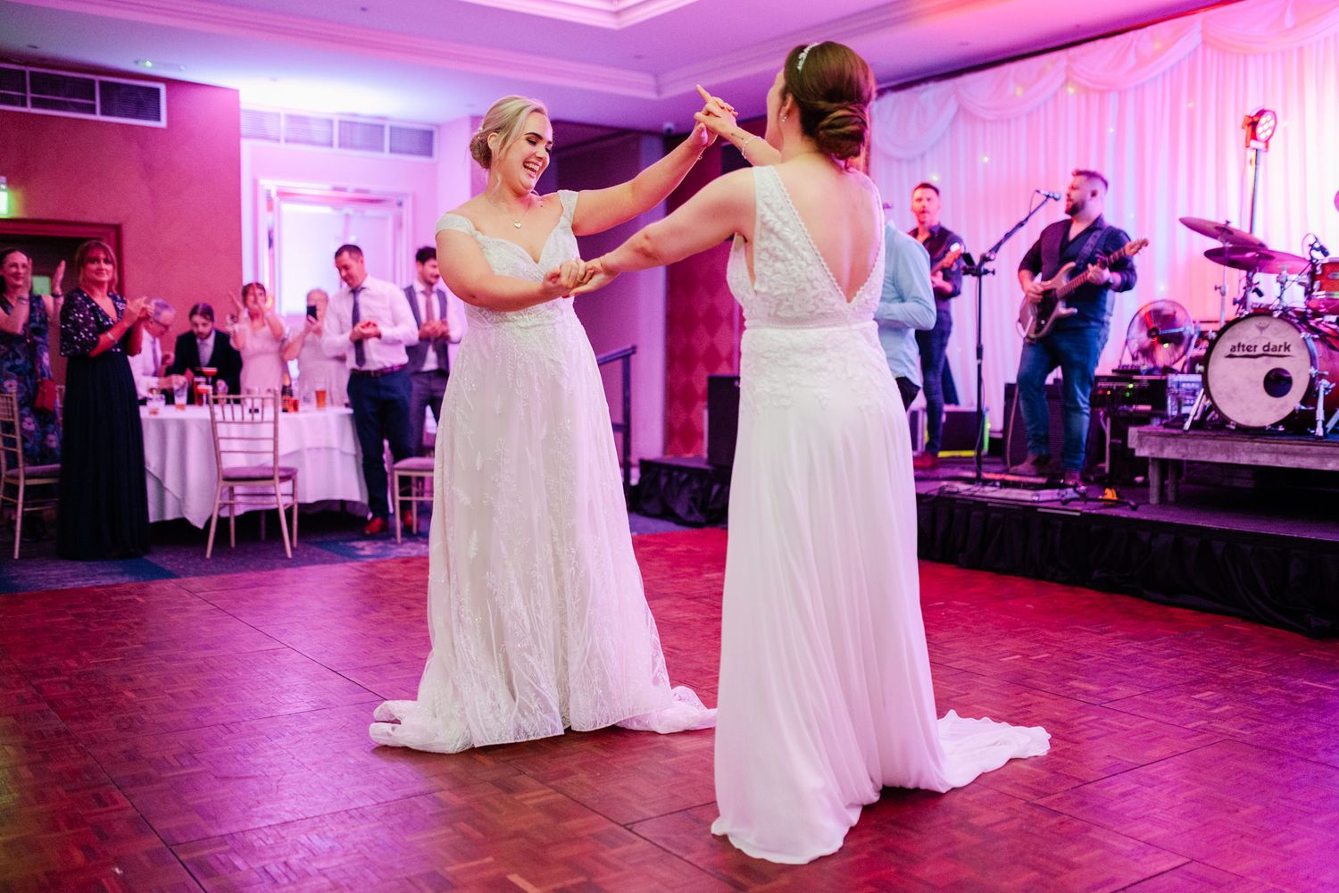 Two people in white dresses dance together in a pink-lit wedding reception venue surrounded by guests.