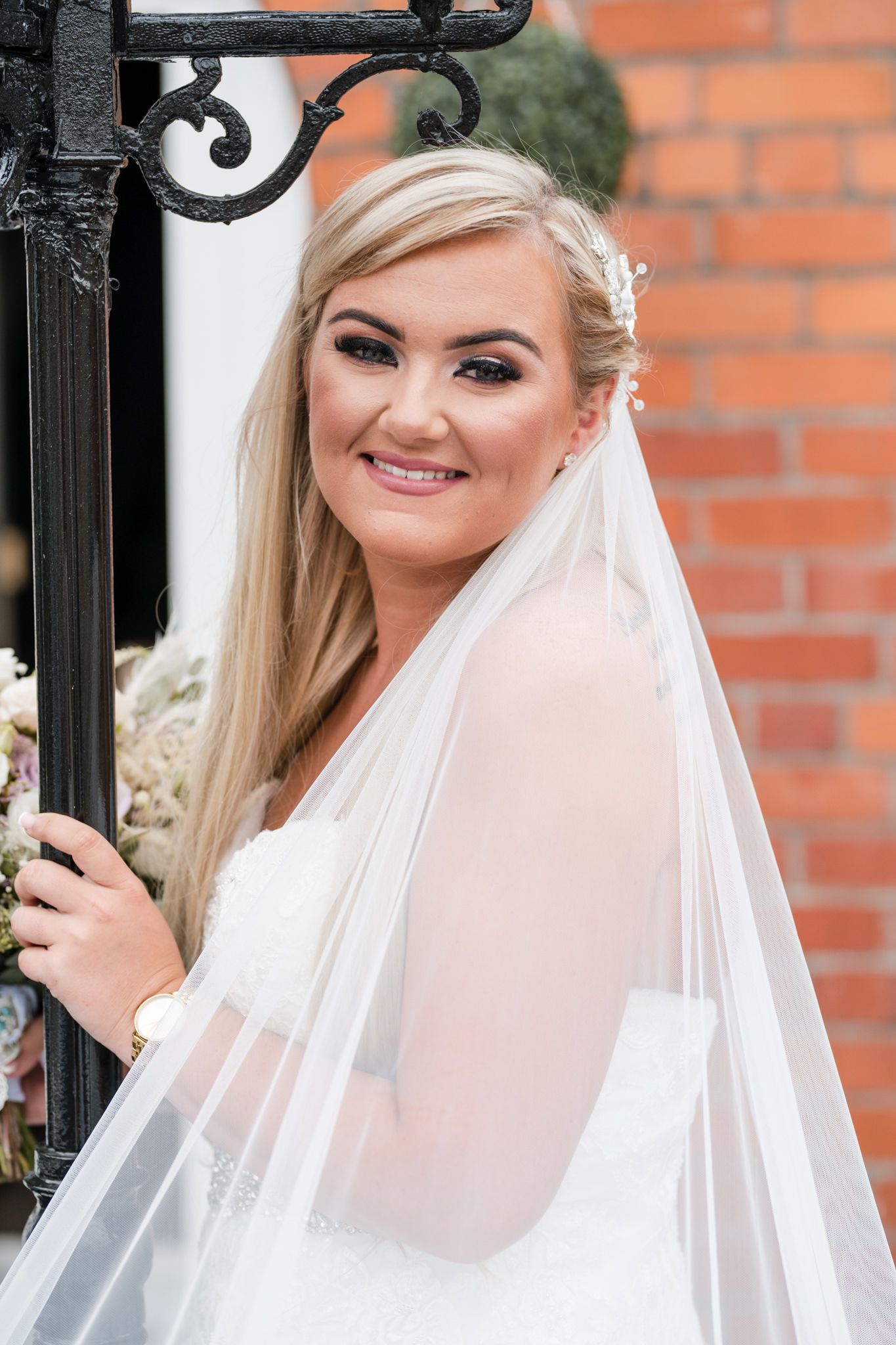 Bride in elegant white wedding dress and veil poses against brick wall backdrop on her special day.