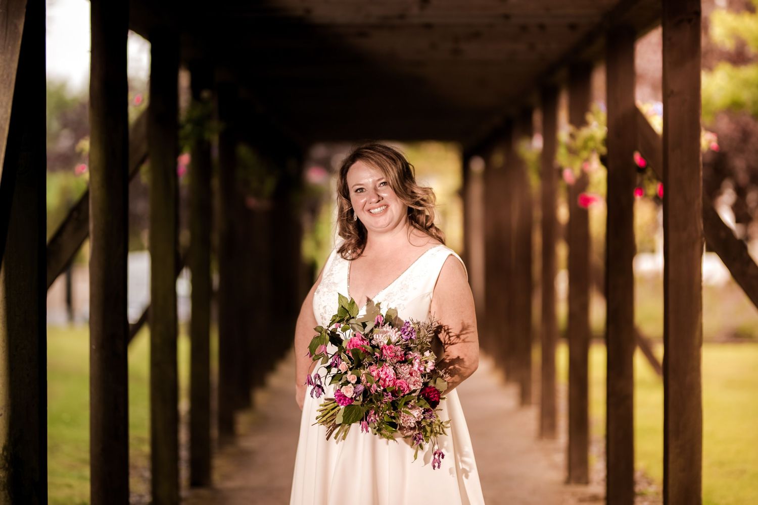 A person in a white wedding dress holds a vibrant pink and red floral bouquet while standing in a covered walkway.