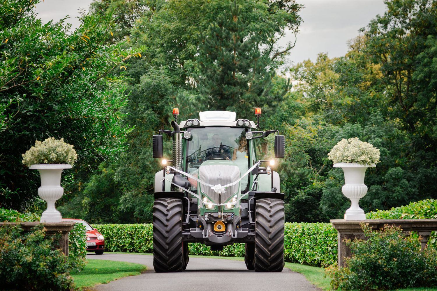 White tractor positioned between decorative garden urns on a formal path surrounded by lush greenery.