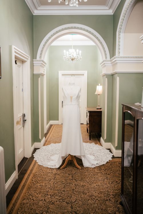 An elegant white wedding dress hangs in a hallway with sage green walls, ornate archways and a vintage chandelier.