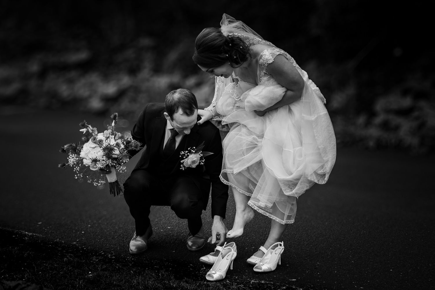Black and white wedding photo showing a playful moment with bouquets outdoors.