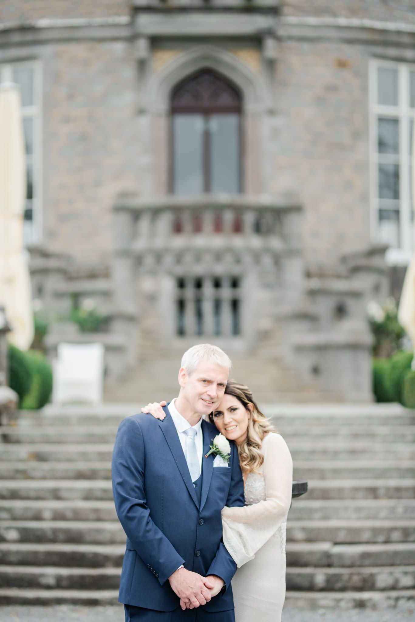 A couple embraces on the grand steps of a stone building with gothic architectural details.