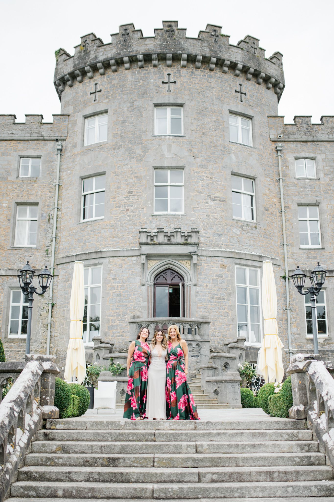 Wedding party standing on stone steps in front of a grand castle-style building with crenellated tower and white windows.