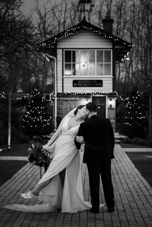 A romantic newlywed couple share a private moment outside a quaint house decorated with twinkling Christmas lights.
