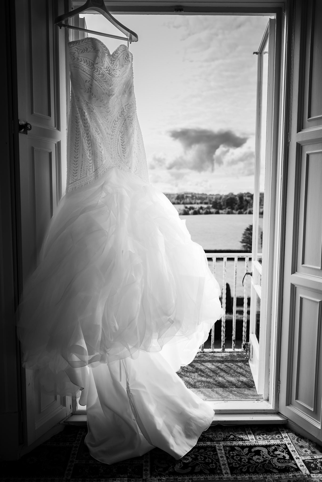 A white wedding dress with sweetheart neckline and tulle skirt hangs in a doorway overlooking a scenic balcony view.