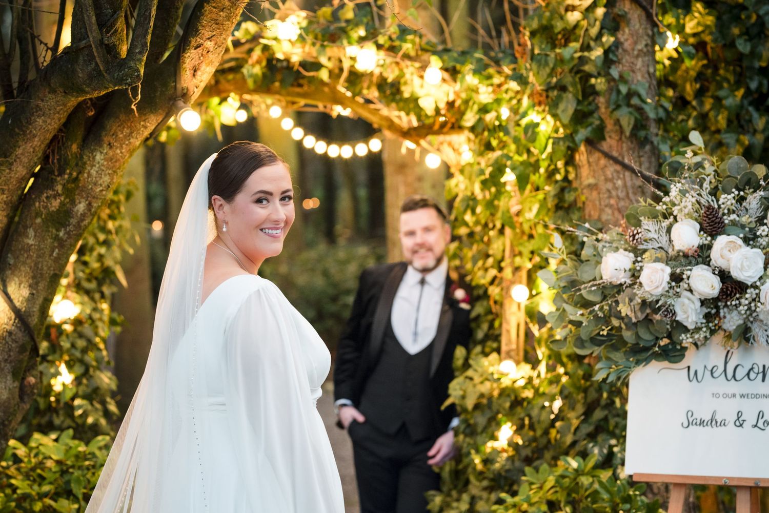 Romantic outdoor wedding ceremony scene under illuminated floral arch at dusk with welcome sign decoration.
