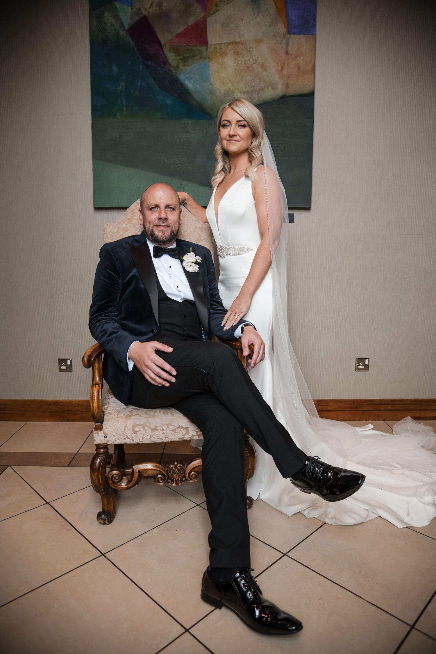 Formal wedding portrait showing couple in elegant attire with groom seated in ornate chair and bride standing behind.