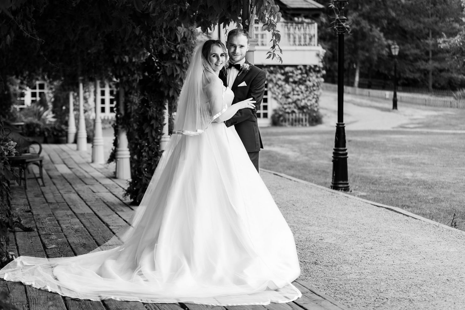 A romantic black and white wedding photo showing a couple standing on a brick walkway by a garden veranda.