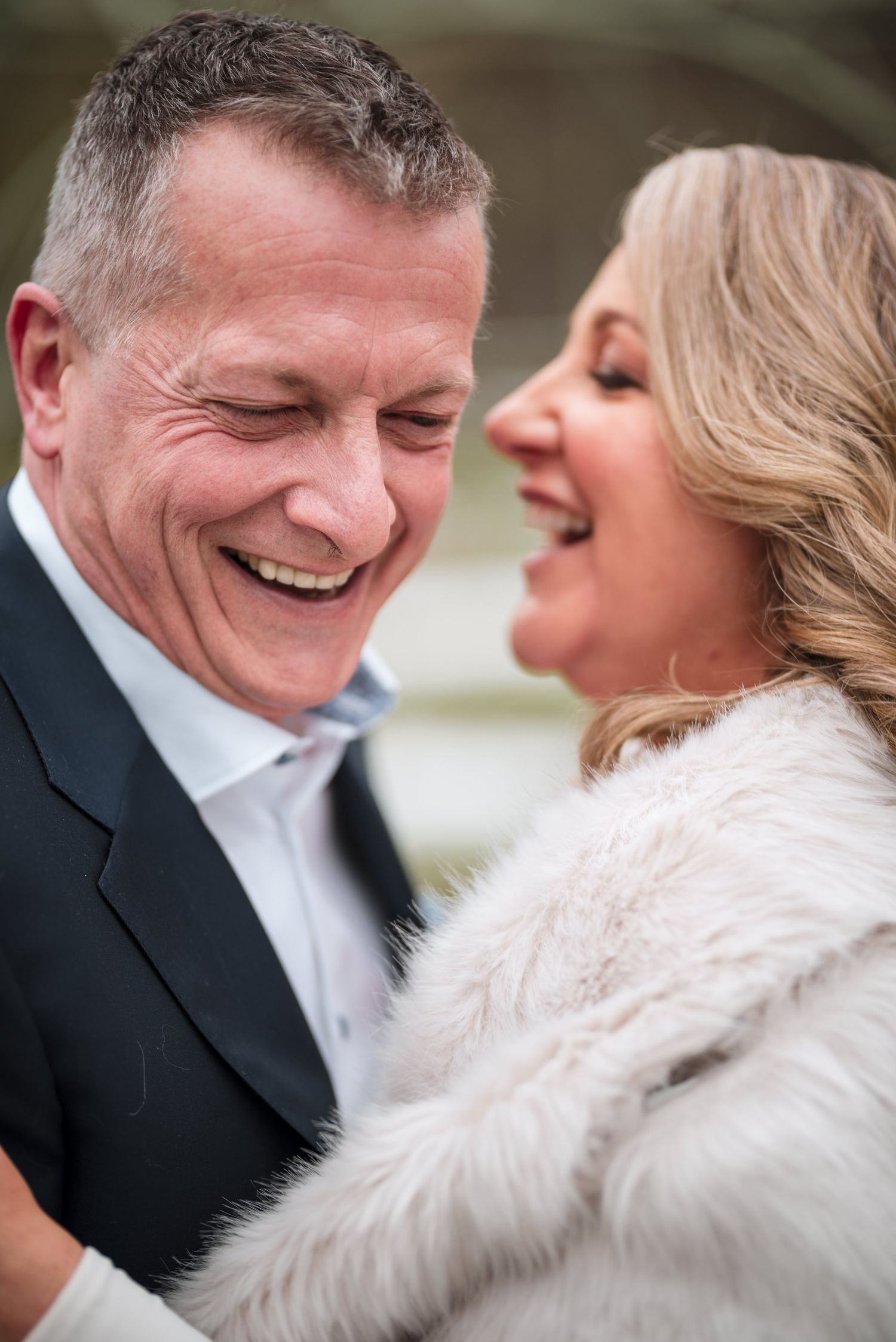 A couple sharing a joyful candid moment while dressed formally, with the person in white wearing a fuzzy winter wrap.