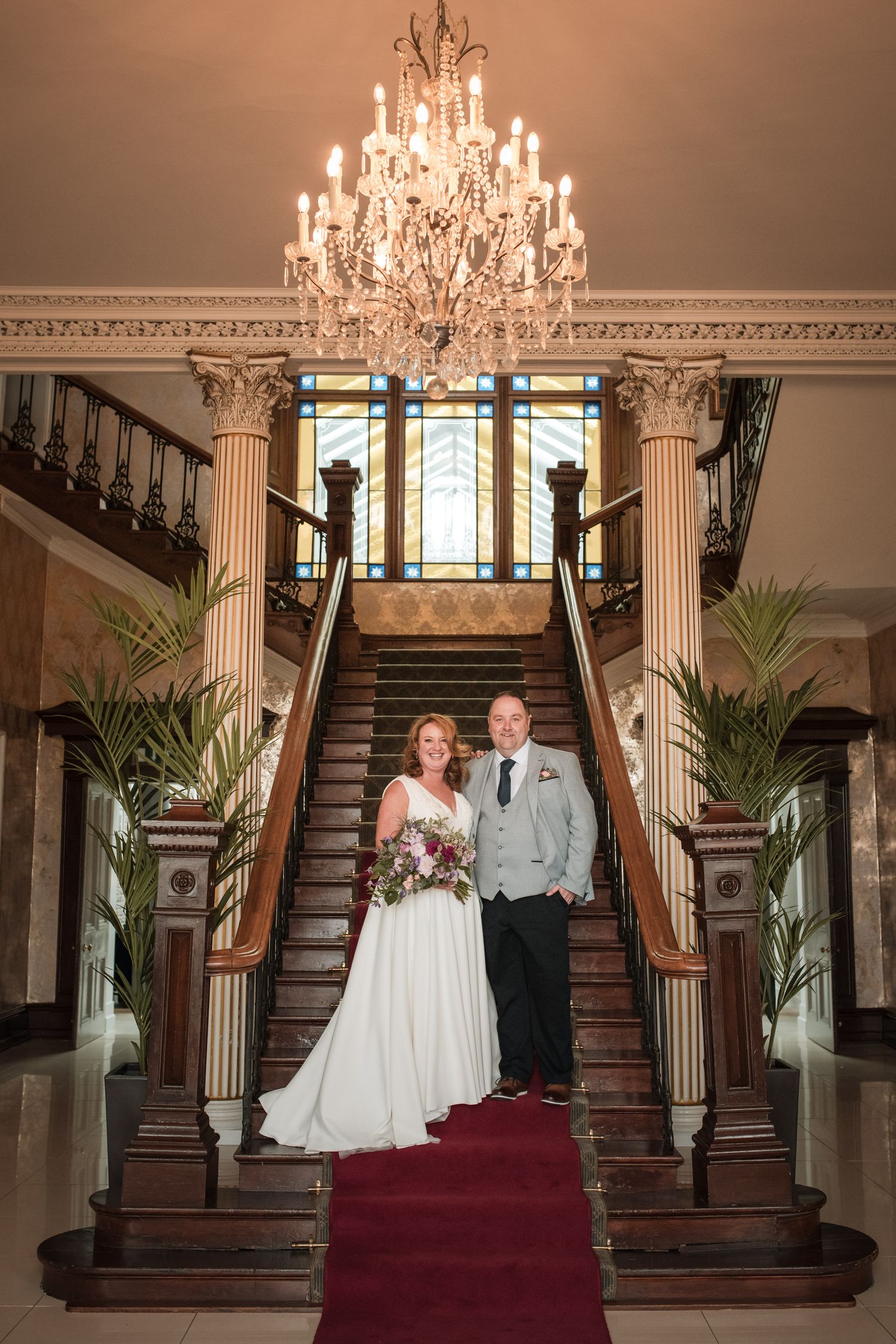 A couple stands on an elegant grand staircase with a crystal chandelier and red carpet in a luxurious historic mansion.