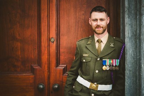 A person wearing military dress uniform with medals stands before a rustic wooden background.