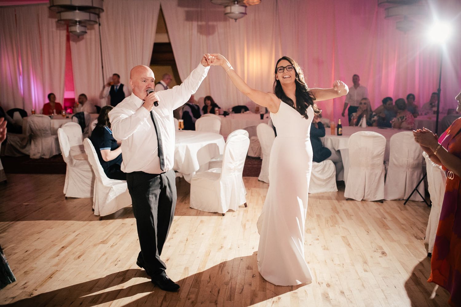 A couple dancing together at a wedding reception in a ballroom with white chairs and pink lighting.