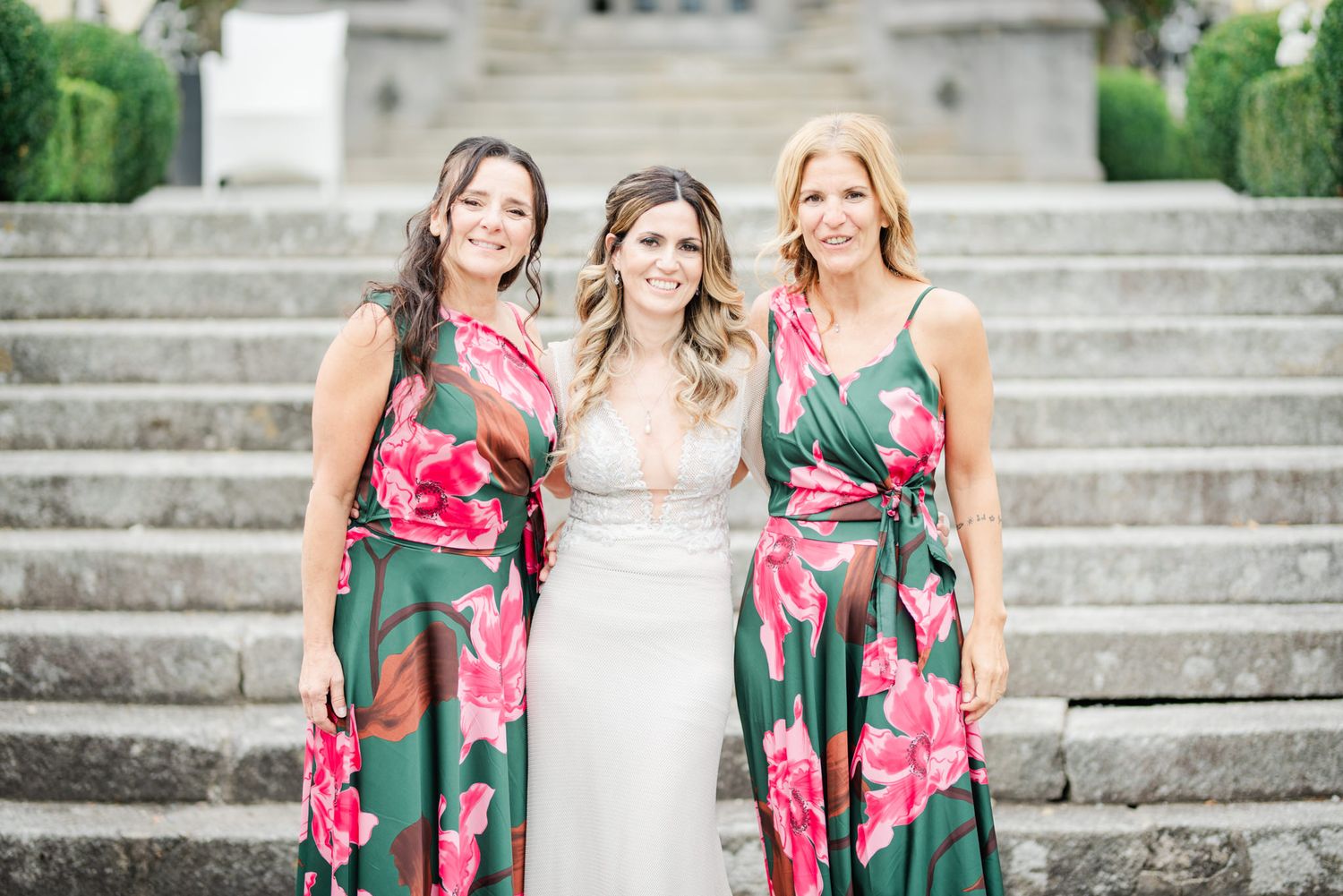 Three people in floral dresses pose on stone steps, with two wearing matching green and pink tropical print gowns.