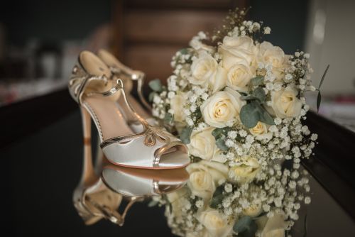 Elegant champagne-colored bridal heels next to a delicate white rose and baby's breath wedding bouquet on reflective surface.