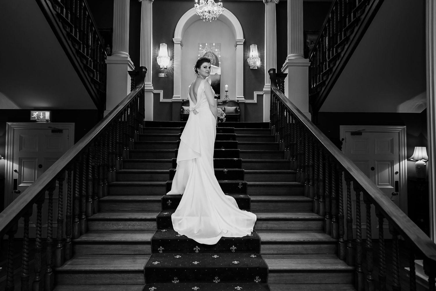 Black and white photograph showing a bride in an elegant lace wedding gown posing on a grand staircase in an ornate venue.