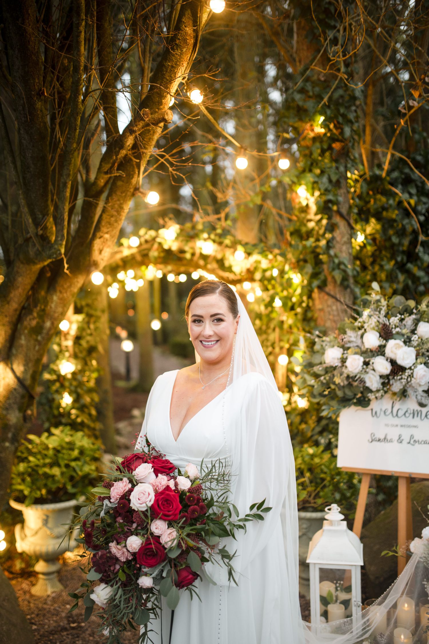 A bride holds a burgundy and pink bouquet while standing in a romantic garden setting with twinkling lights and trees.