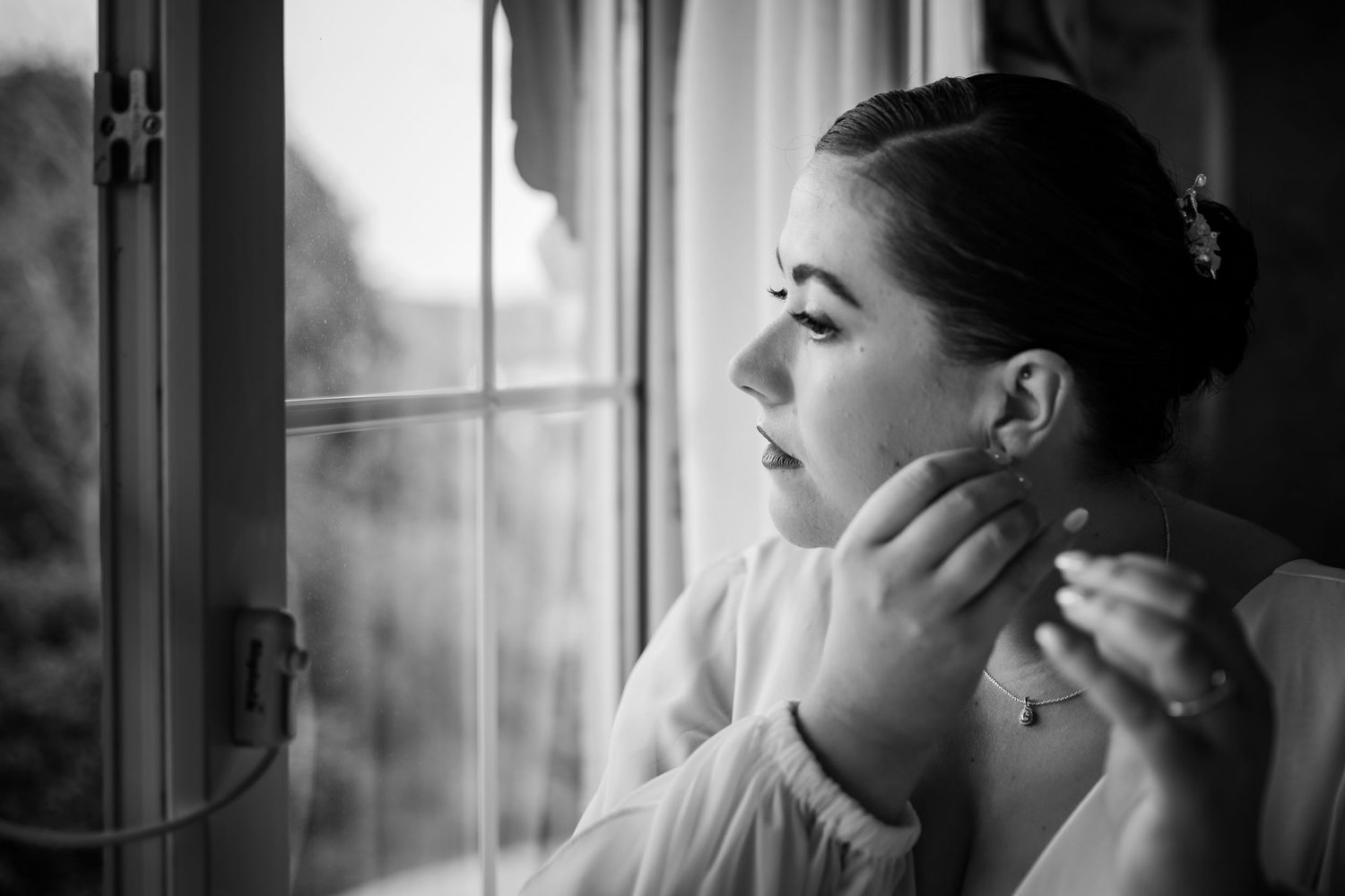Black and white portrait of a woman putting on earrings by a window.