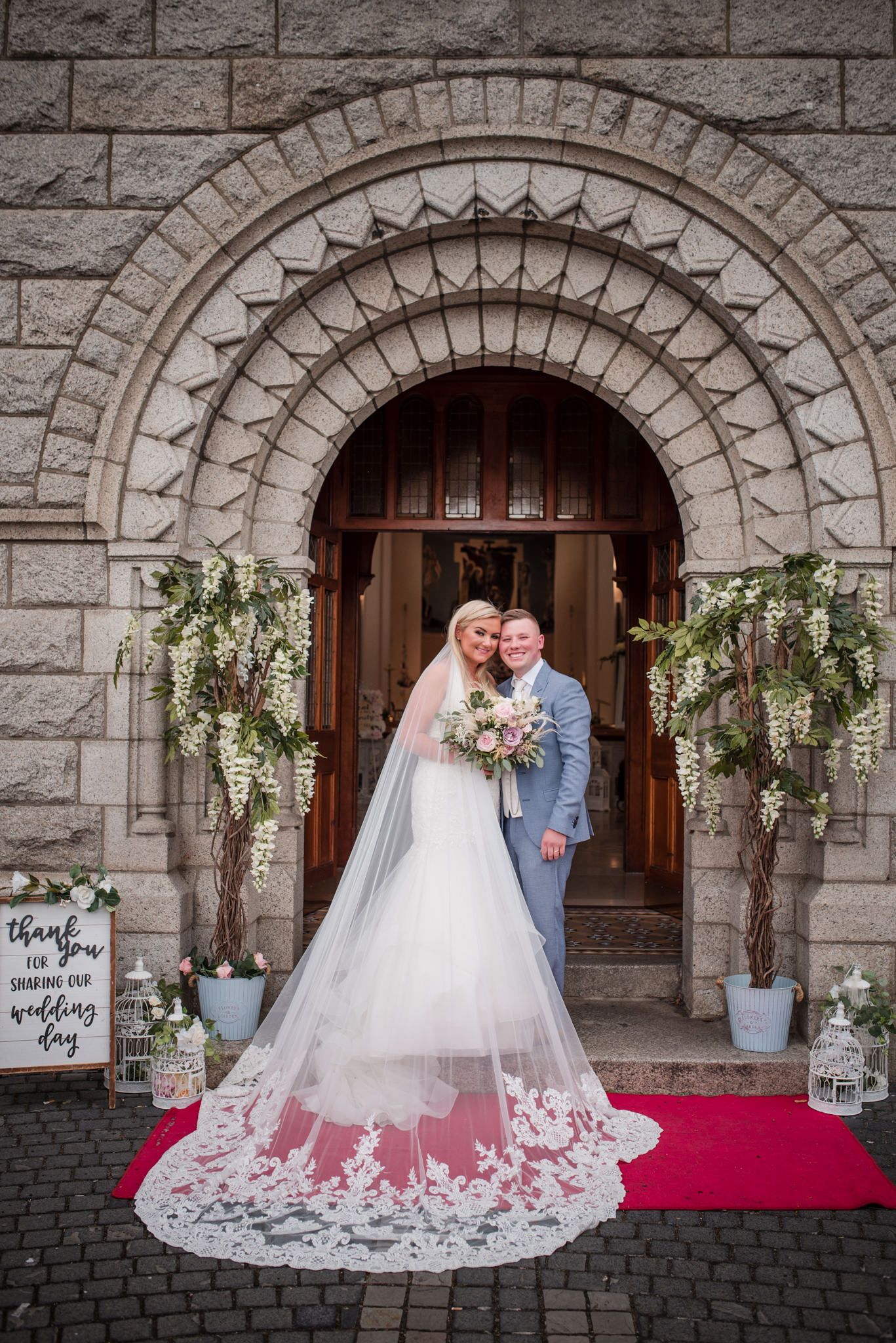 A couple poses in an ornate stone archway entrance with hanging floral decor and a long lace wedding veil on a red carpet.