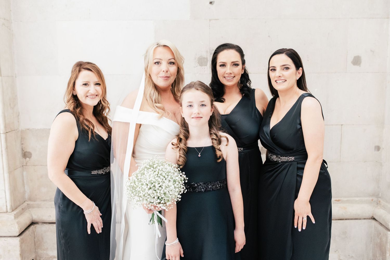Group in elegant black dresses pose together against light wall with one in white gown holding delicate baby's breath bouquet.