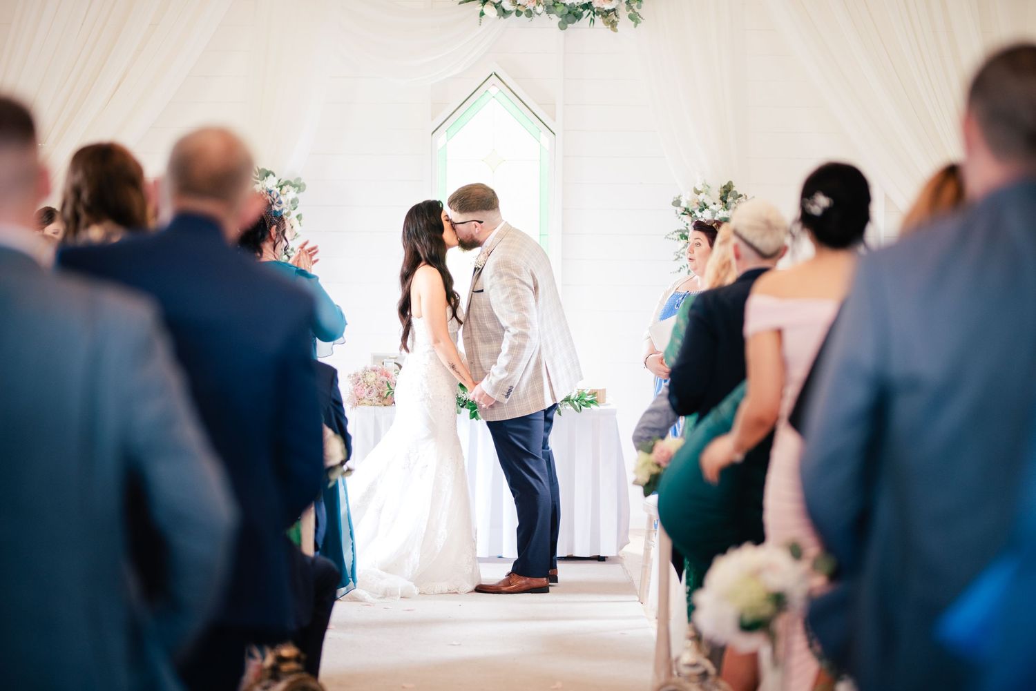 A romantic wedding ceremony takes place inside a bright white chapel with guests watching from their seats.