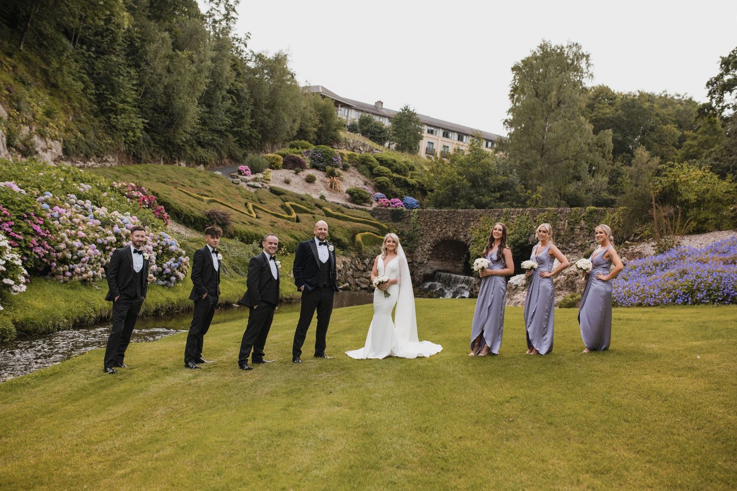 Wedding party members lined up on lawn with purple flowers and greenery in elegant outdoor garden setting.