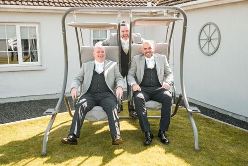 Three people in formal gray suits and vests smile while sitting on an outdoor patio swing in front of a white building.