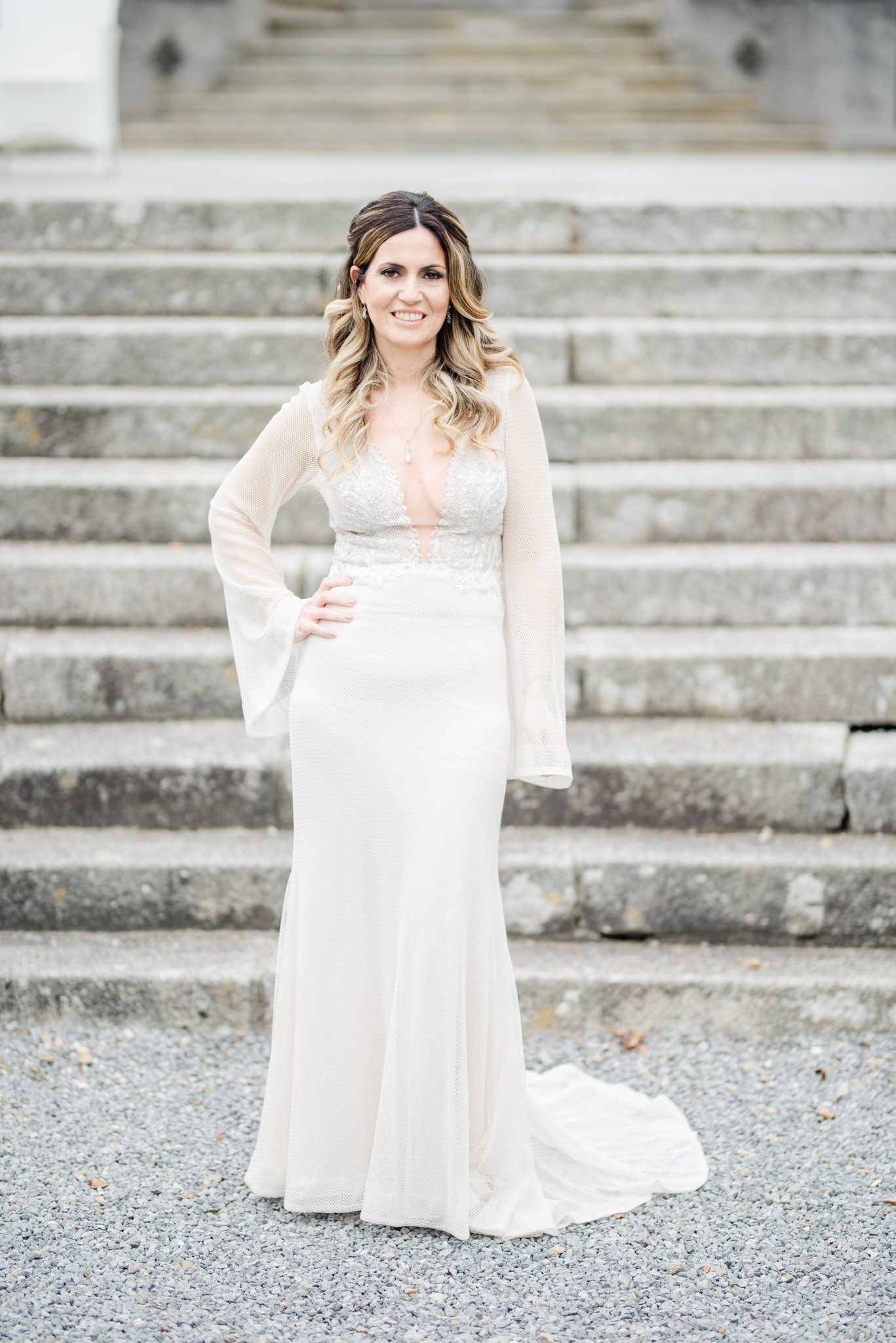 A bride in a flowing white dress with bell sleeves poses elegantly on stone steps at an outdoor venue.