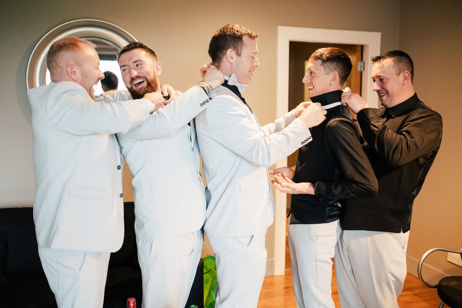 A group of people in light colored outfits help adjust each other's clothes while getting ready in a room.
