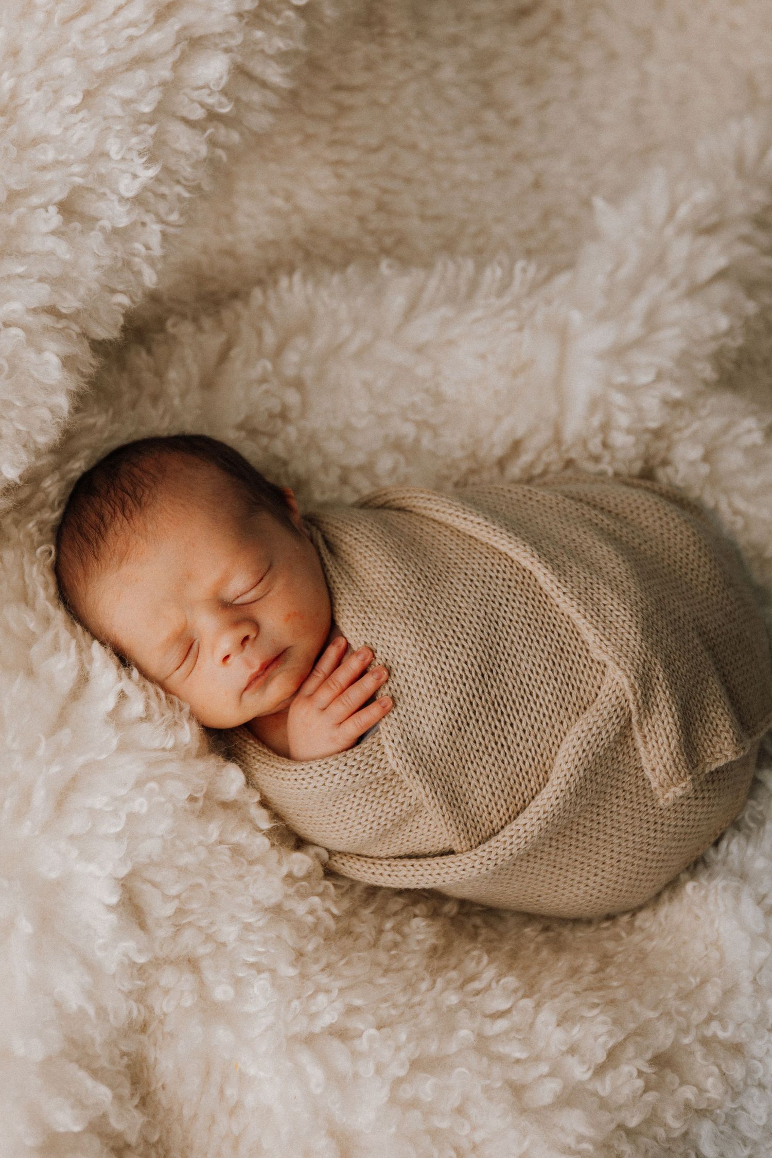 A sleeping newborn baby wrapped in a beige knit blanket lies on soft white fur in a series of peaceful poses.