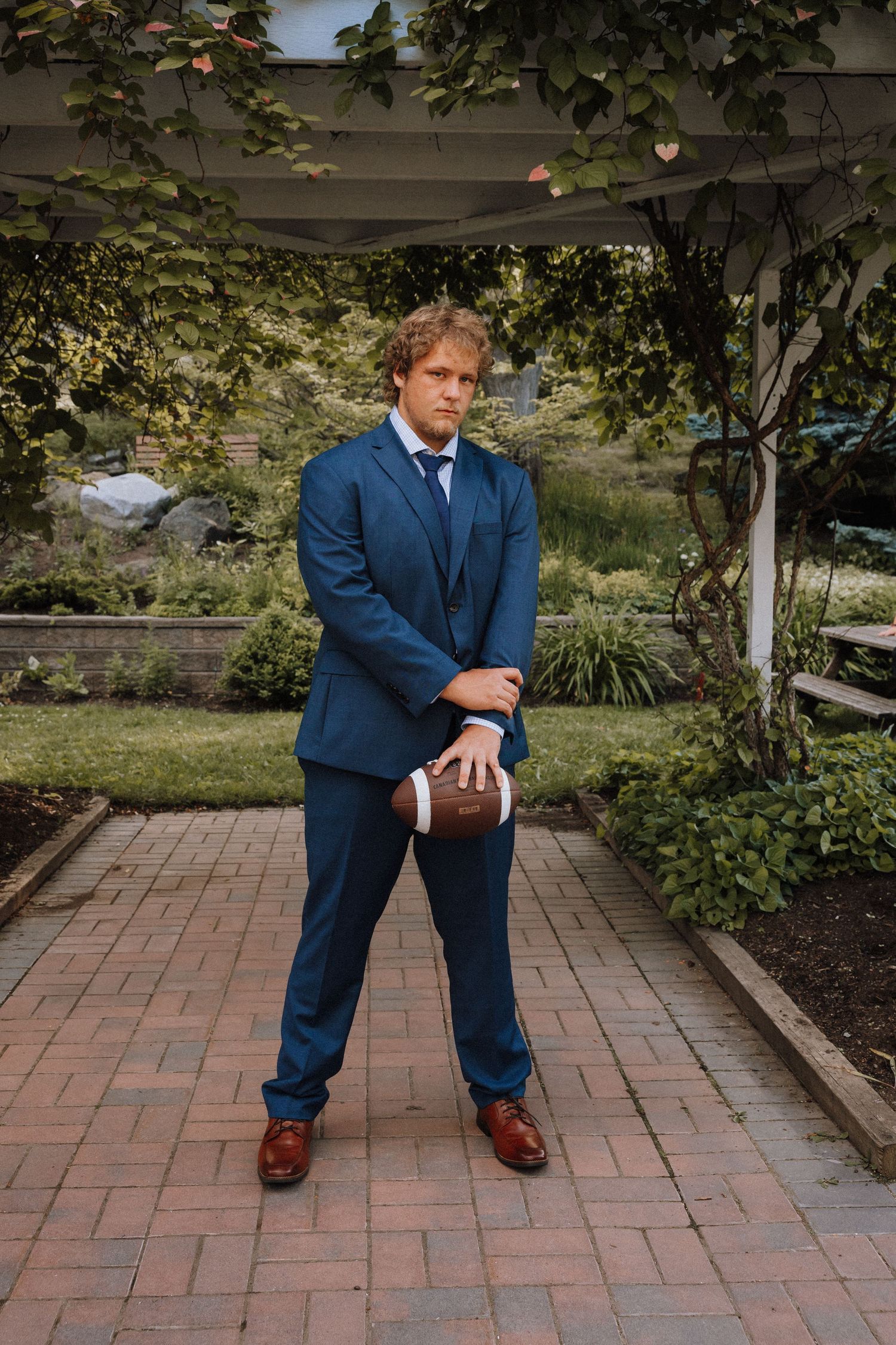 Man in blue suit holding football on brick path in lush garden under white pergola.