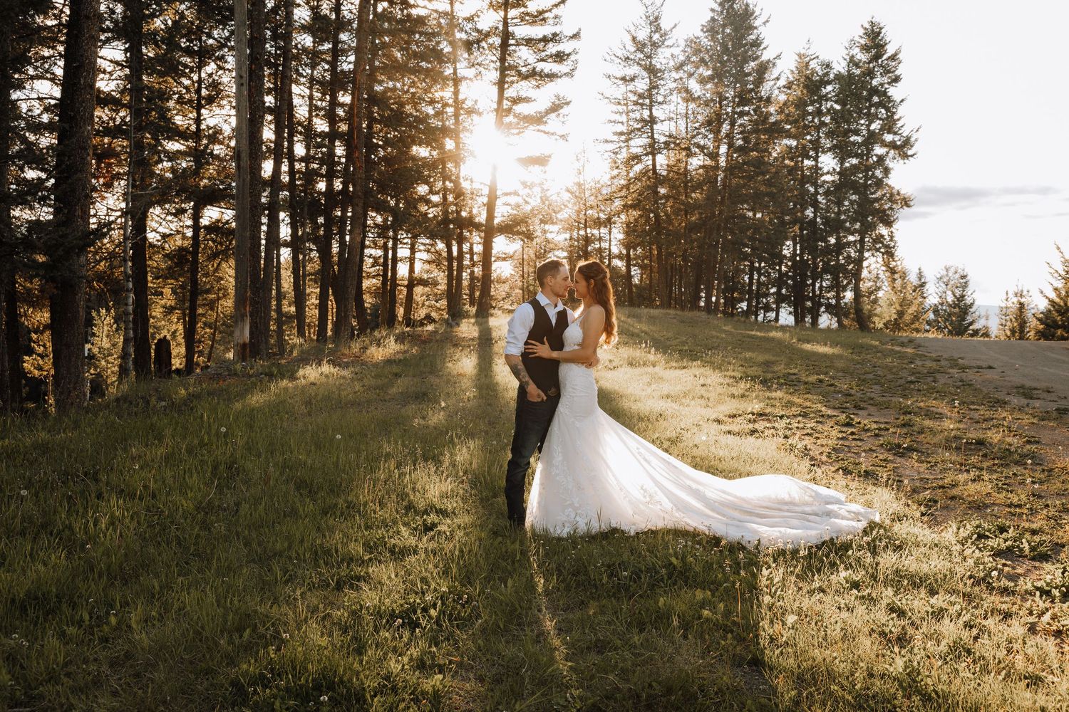 Bride and groom share a romantic kiss in a sunlit forest meadow, with golden light streaming through tall pine trees.