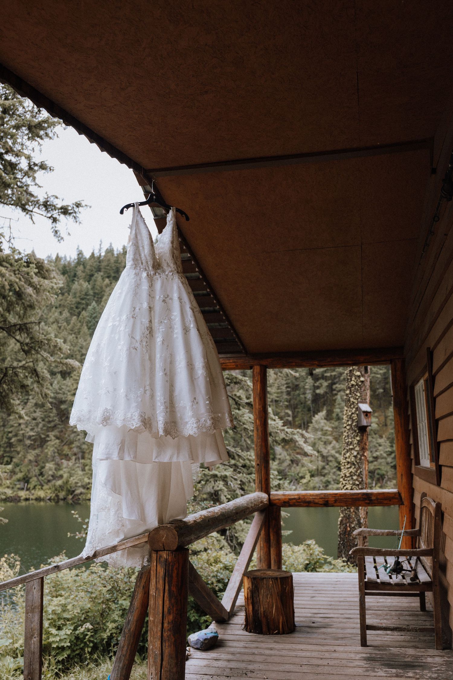 A delicate white lace wedding dress hangs on display on a rustic cabin porch with a lake view in the background.